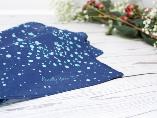 furoshiki or scarf laid on table. Navy blue pattern with stars and green accent. A Kindly Yarn logo is printed on the edge of the fabric